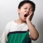 Child experiencing tooth pain, holding cheek with hand and yawning widely.