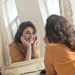 Woman in yellow shirt smiling at mirror, demonstrating proper dental care routine.
