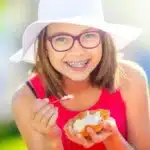 Girl with braces enjoying a summer snack outdoors while wearing glasses and a white hat.