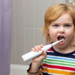 Young child brushing teeth with electric toothbrush in bathroom.