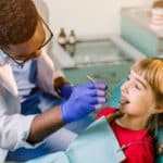 Pediatric dentist examining young girl's teeth in dental chair during checkup.
