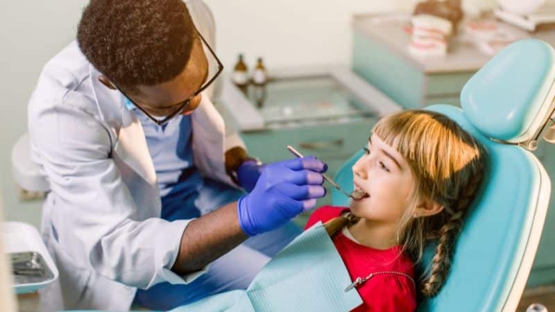 Pediatric dentist examining young girl's teeth in dental chair during checkup.