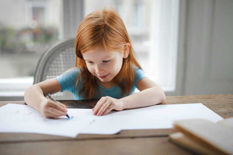 Young girl with red hair concentrating while writing at a desk, illustrating student focus and academic engagement.