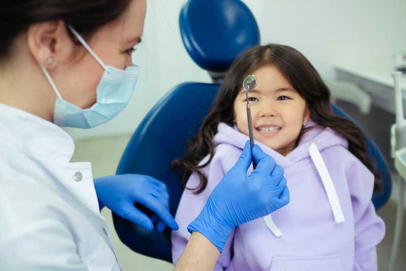 Young girl smiling in dental chair while pediatric dentist examines her teeth with dental mirror.