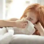 Child with red hair blowing nose into tissue while sick in bed.