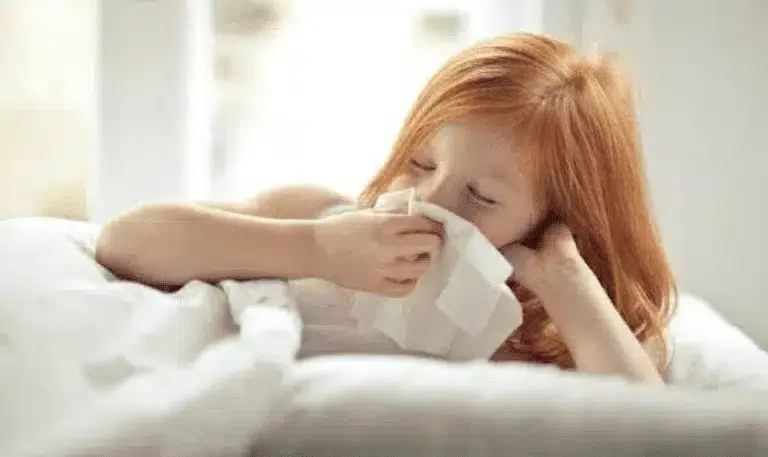 Child with red hair blowing nose into tissue while sick in bed.