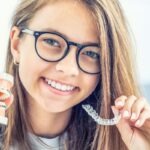 Teen girl with glasses smiling while holding dental model and clear aligner tray.