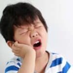 Child experiencing tooth pain, holding cheek with closed eyes and open mouth.