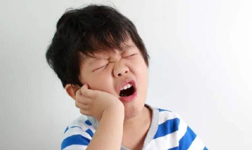Child experiencing tooth pain, holding cheek with closed eyes and open mouth.
