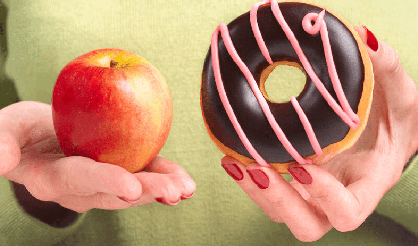 Hand holding a red apple next to a chocolate donut, illustrating healthy versus sugary food choices for children's dental he.