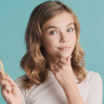 Woman holding a wooden toothbrush against a blue background, considering dental care choices.