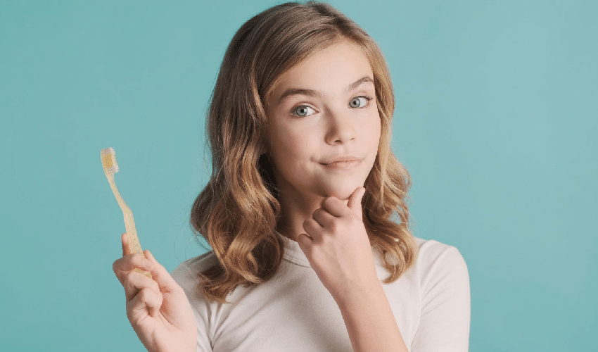 Woman holding a wooden toothbrush against a blue background, considering dental care choices.