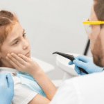 Young girl in dental chair holding her cheek while dentist examines her teeth during emergency visit.