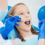 Young girl receiving pediatric dental examination with dentist using tools and mirror.