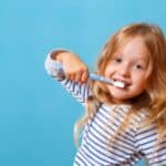 Happy child brushing teeth with blue toothbrush against blue background.