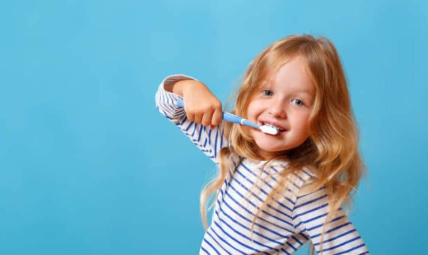Happy child brushing teeth with blue toothbrush against blue background.