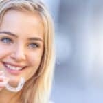 Young woman holding clear orthodontic aligner, smiling brightly at camera.
