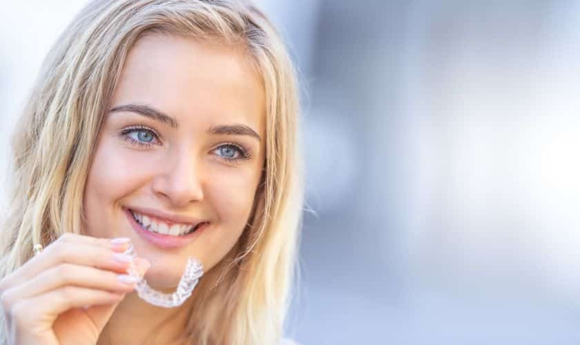 Young woman holding clear orthodontic aligner, smiling brightly at camera.