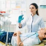 Pediatric dentist in blue gloves explaining sedation options to young patient in dental chair.