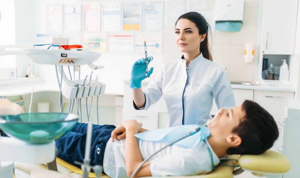 Pediatric dentist in blue gloves explaining sedation options to young patient in dental chair.
