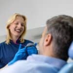 Smiling female dentist in blue scrubs discussing treatment with patient in dental chair.