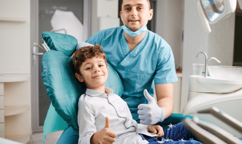 Pediatric dentist with young patient giving thumbs up in dental office treatment room.