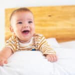 Happy baby in yellow striped shirt smiling on bed, illustrating positive childhood development during teething stage.
