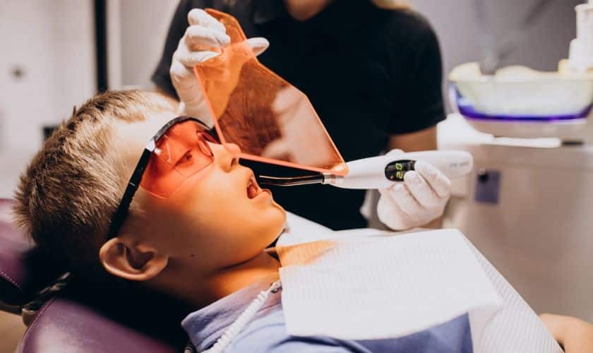 Young patient wearing protective eyewear during laser dental treatment with dentist holding instrument.