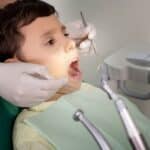 Young child receiving dental examination in orthodontic chair with dentist wearing gloves and using dental instruments.