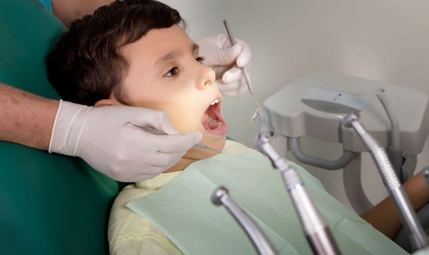 Young child receiving dental examination in orthodontic chair with dentist wearing gloves and using dental instruments.