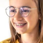Smiling teen girl wearing glasses and yellow shirt displays metal braces on teeth.