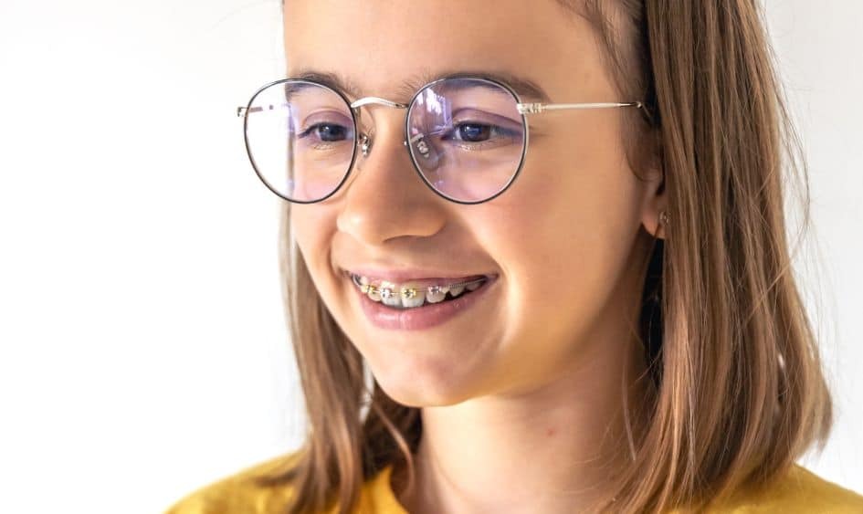 Smiling teen girl wearing glasses and yellow shirt displays metal braces on teeth.
