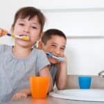 Two young children brushing their teeth at a bathroom sink with colorful toothbrushes.