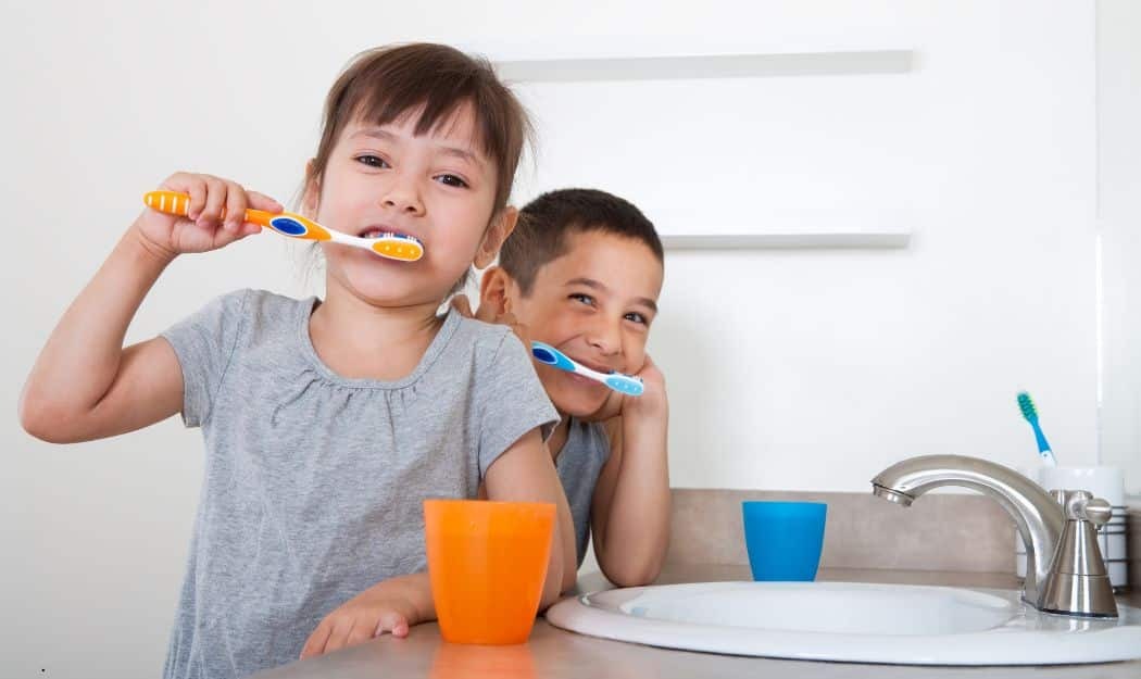 Two young children brushing their teeth at a bathroom sink with colorful toothbrushes.