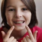 Child with metal braces smiling and pointing to teeth in Spring, TX dental practice.