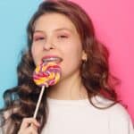 Young girl with braces enjoying a colorful lollipop against a blue and pink gradient background.