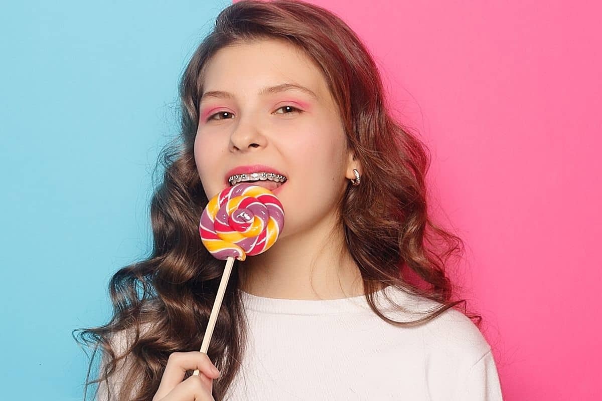 Young girl with braces enjoying a colorful lollipop against a blue and pink gradient background.
