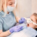 Pediatric dentist examining young patient's teeth during dental procedure with protective eyewear.