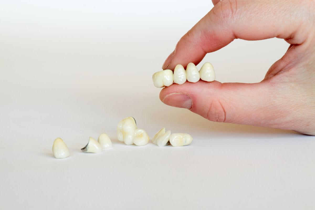 Hand holding dental crown next to scattered crowns on white surface.