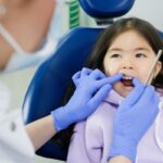 Young girl in dental chair receiving oral examination from dentist wearing blue gloves.