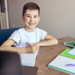 Smiling boy with braces doing homework at desk with notebooks and pencils.