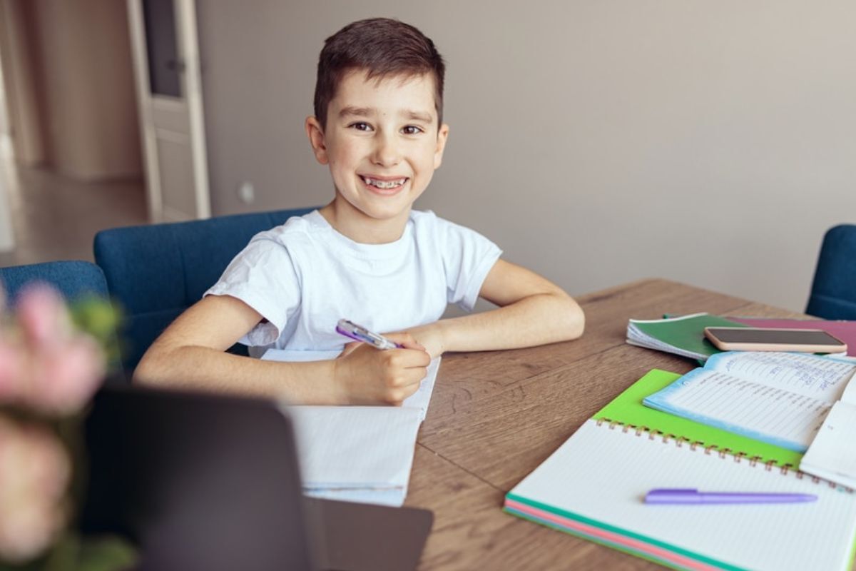 Smiling boy with braces doing homework at desk with notebooks and pencils.