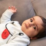 Infant in white outfit with red heart detail lying on gray couch.