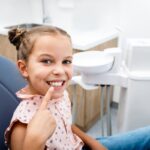Happy young girl in dental chair pointing to her healthy smile during pediatric dental visit.