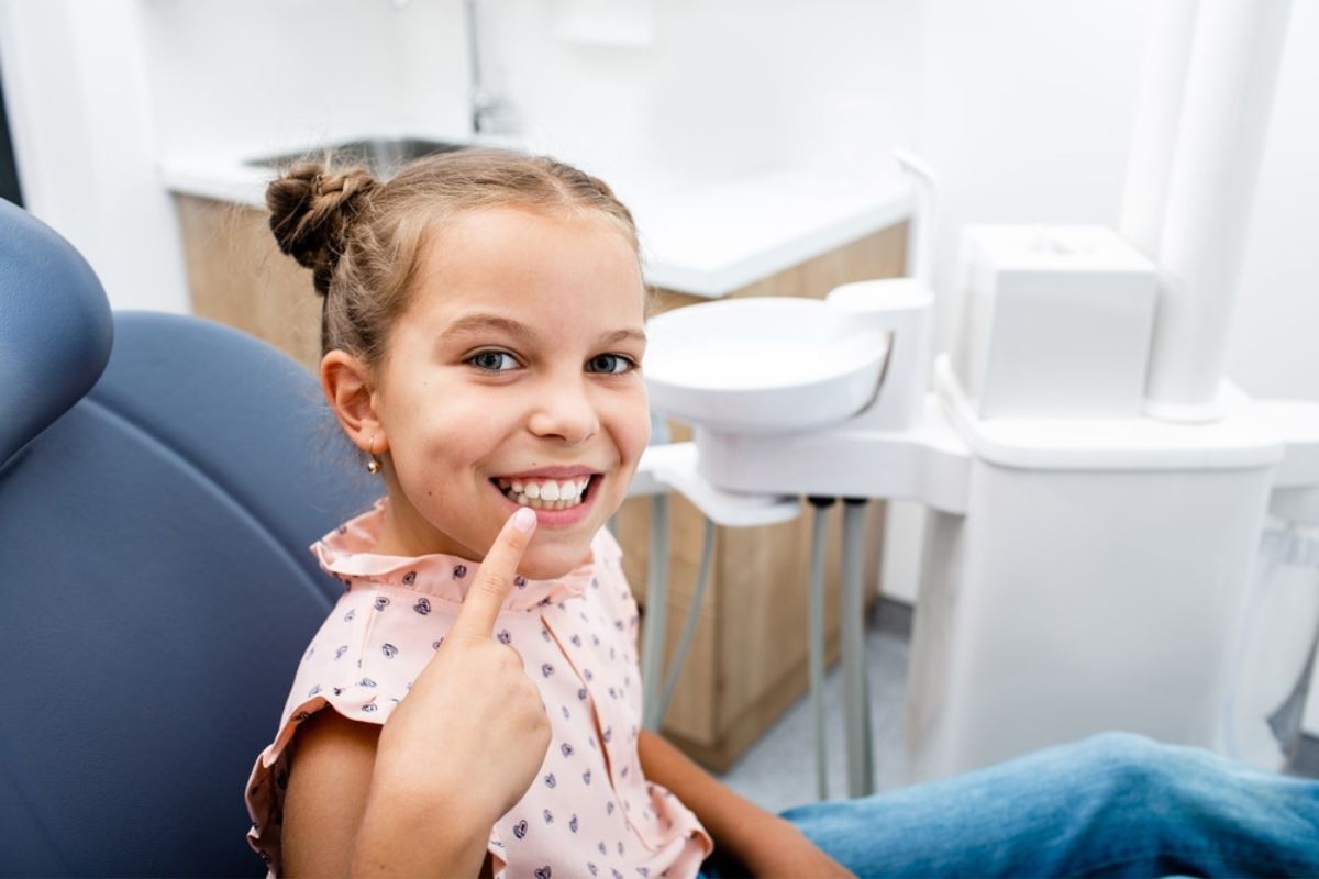 Happy young girl in dental chair pointing to her healthy smile during pediatric dental visit.