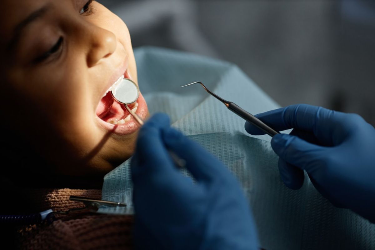 Pediatric dentist in blue gloves examining child's teeth with dental mirror and tools.