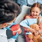 Young girl smiling with teddy bear during pediatric dental visit with dentist holding teeth model.