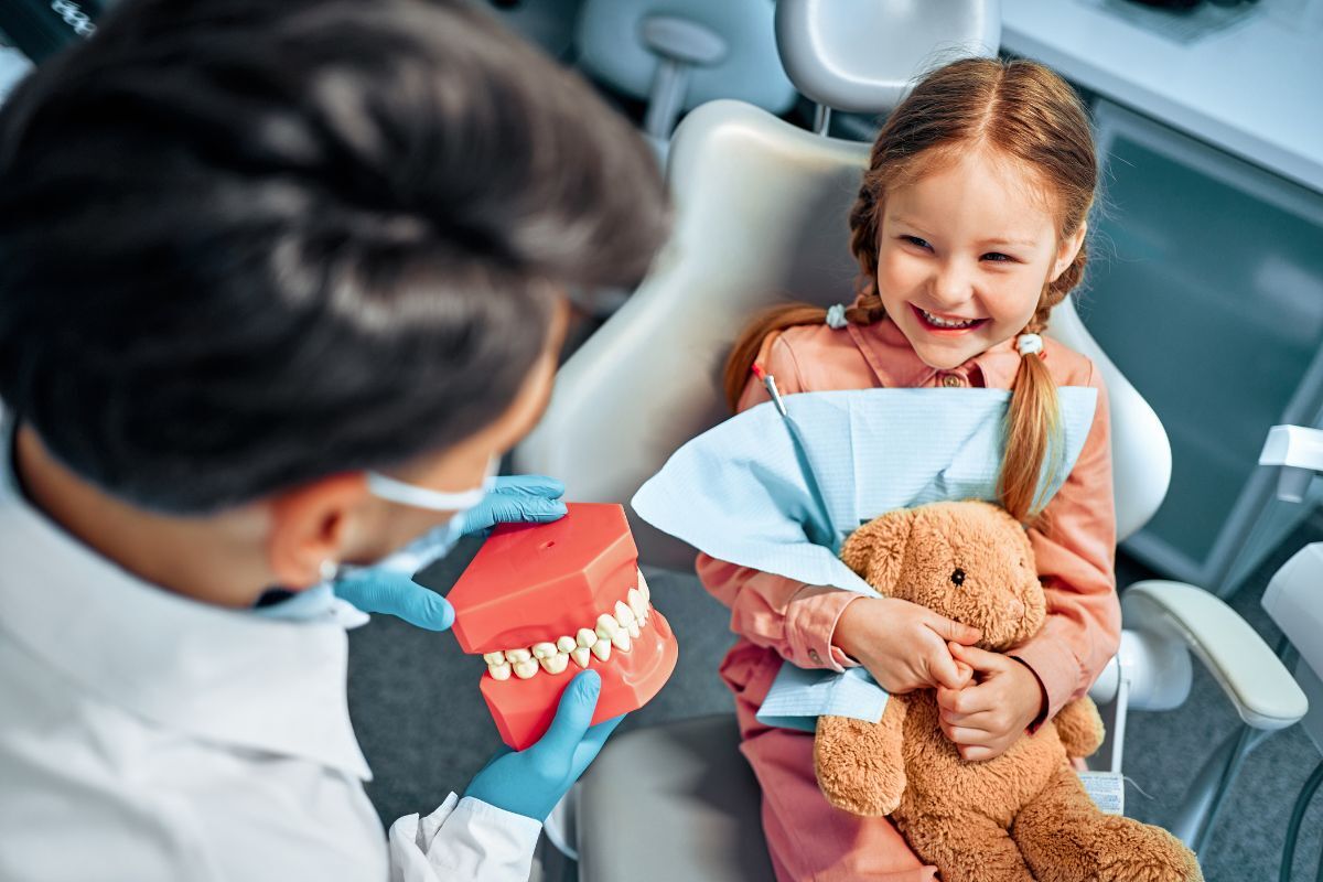 Young girl smiling with teddy bear during pediatric dental visit with dentist holding teeth model.