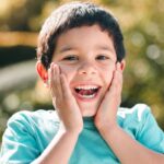 Young boy in turquoise shirt smiling with hands to face, expressing excitement about dental care.