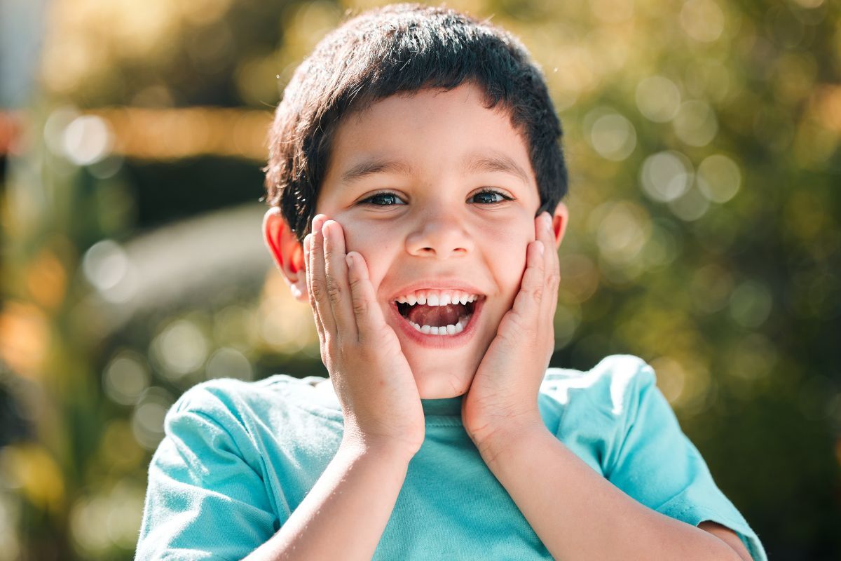 Young boy in turquoise shirt smiling with hands to face, expressing excitement about dental care.